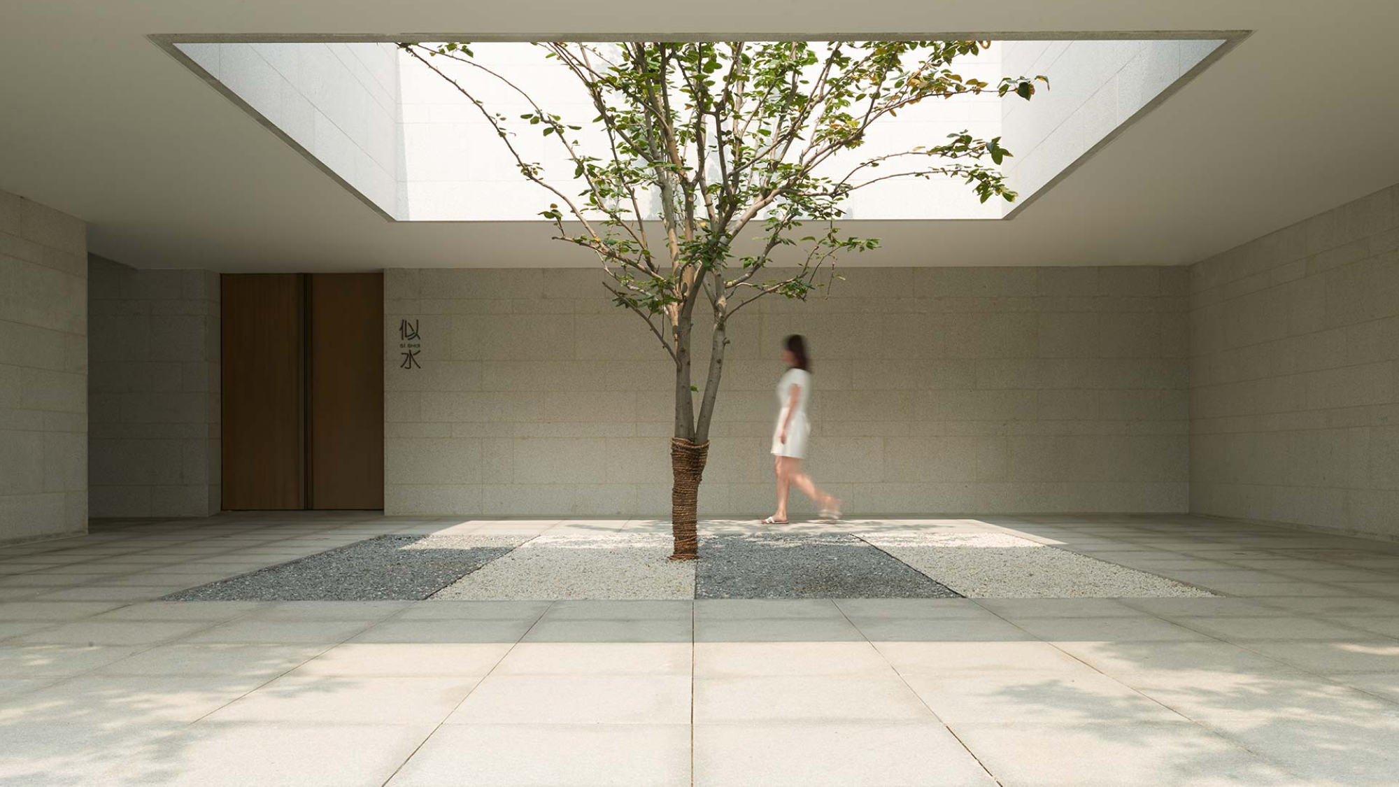 tree in the middle of a courtyard with an opening in the rood and woman walking 