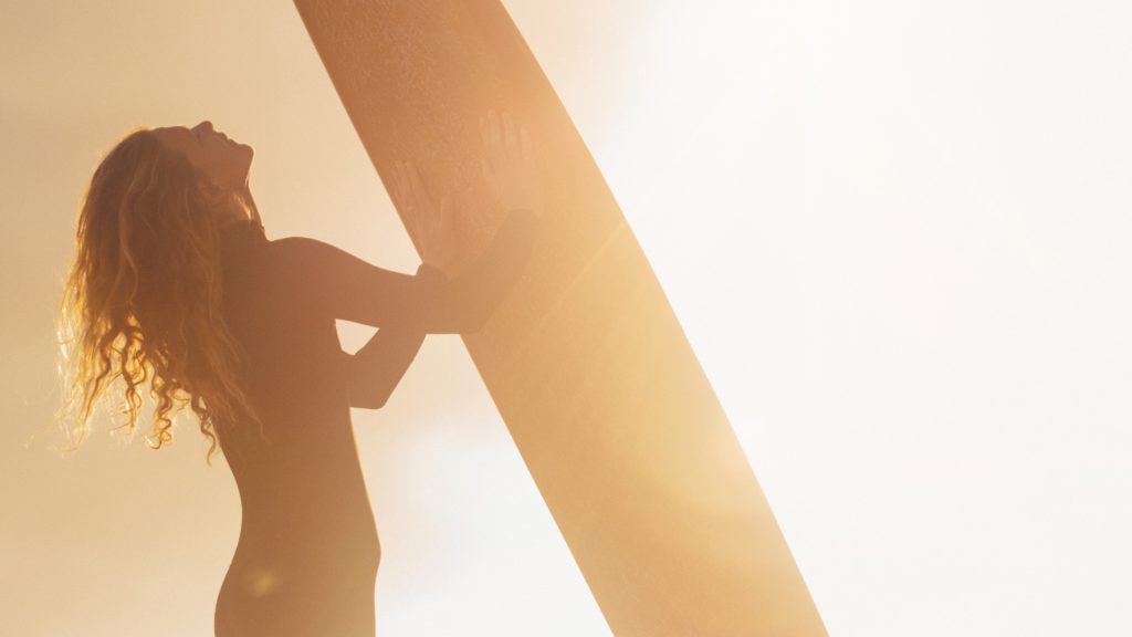 woman holding a surf board looking up to the sun