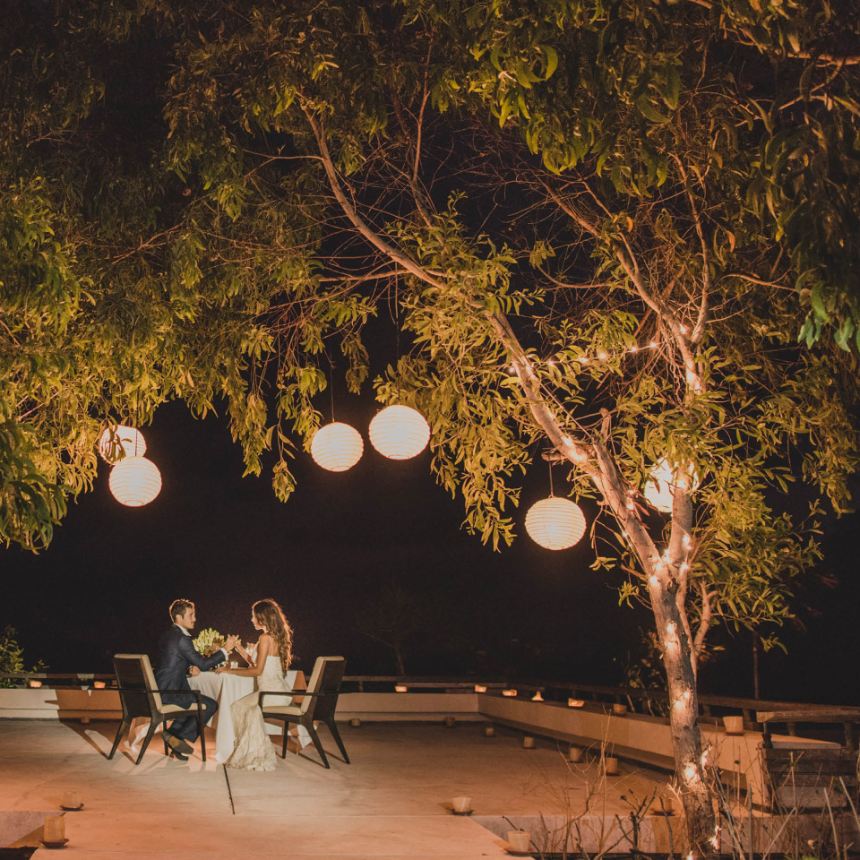 Wedding couple sitting at a table under lush trees with paper lanters.