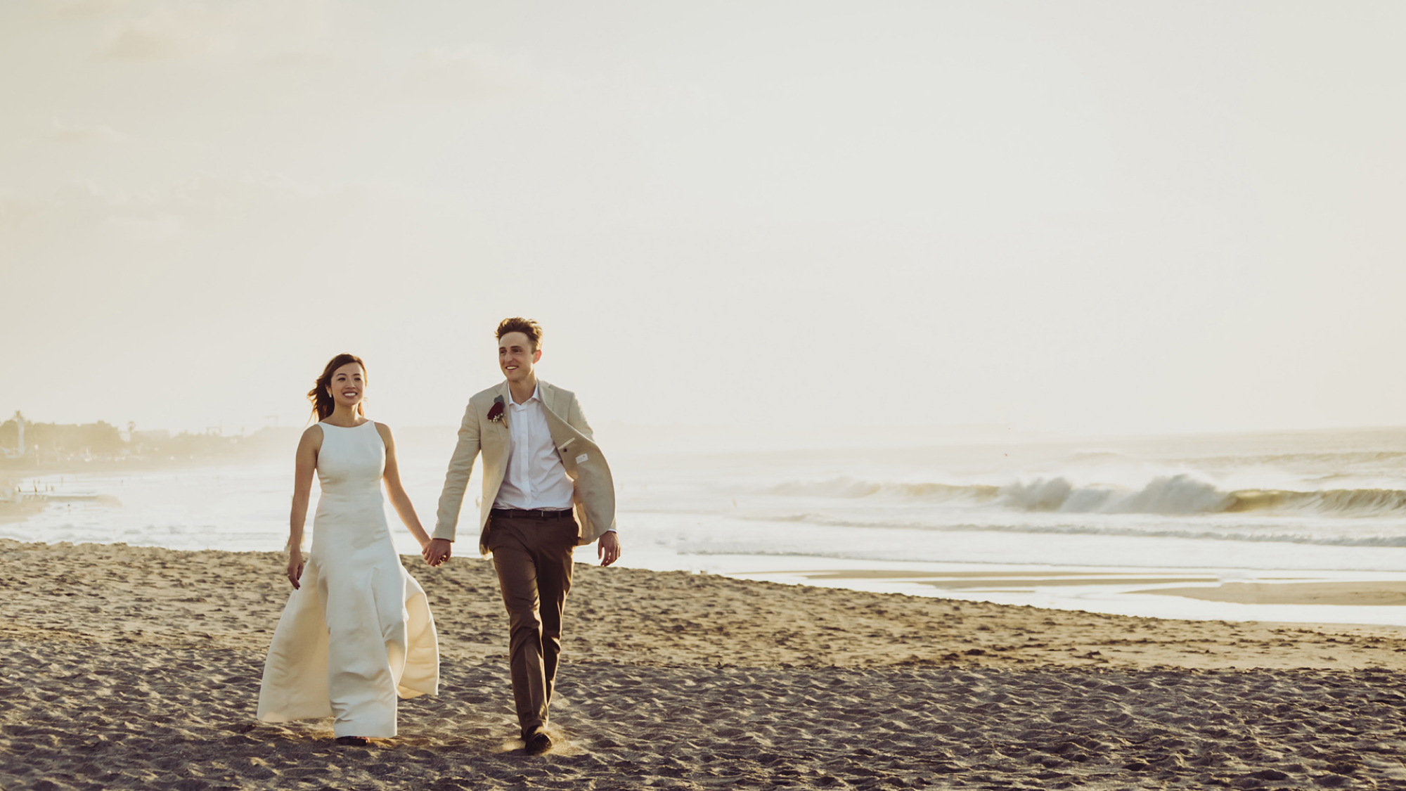 A bride and groom lookin happy and holding hands while walking on a beach