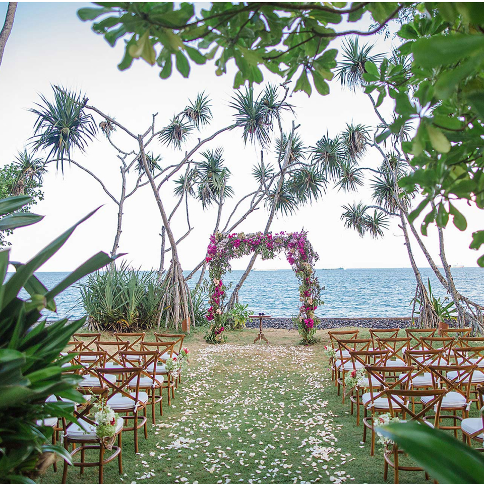 an aisle of flowers leading to a wedding arch wrapped in pink flowers, surrounded by palm trees with an ocean view