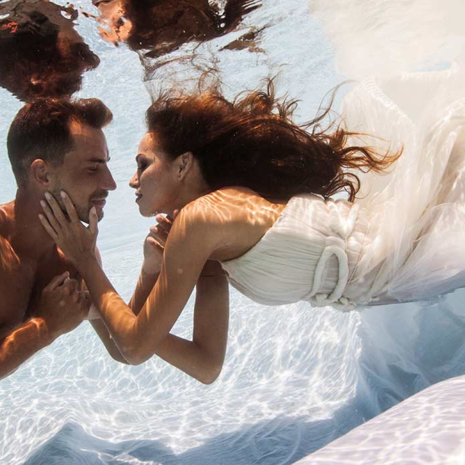 Groom and bride in her white gown looking at each other underwater