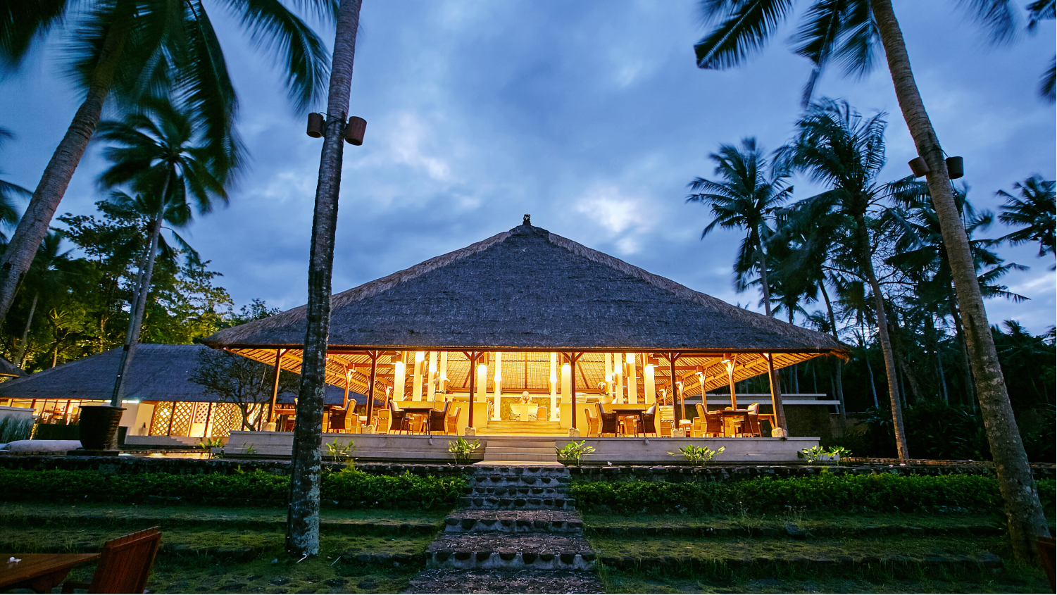 A beautifully lit building with a straw roof surrounded by grass and palm trees