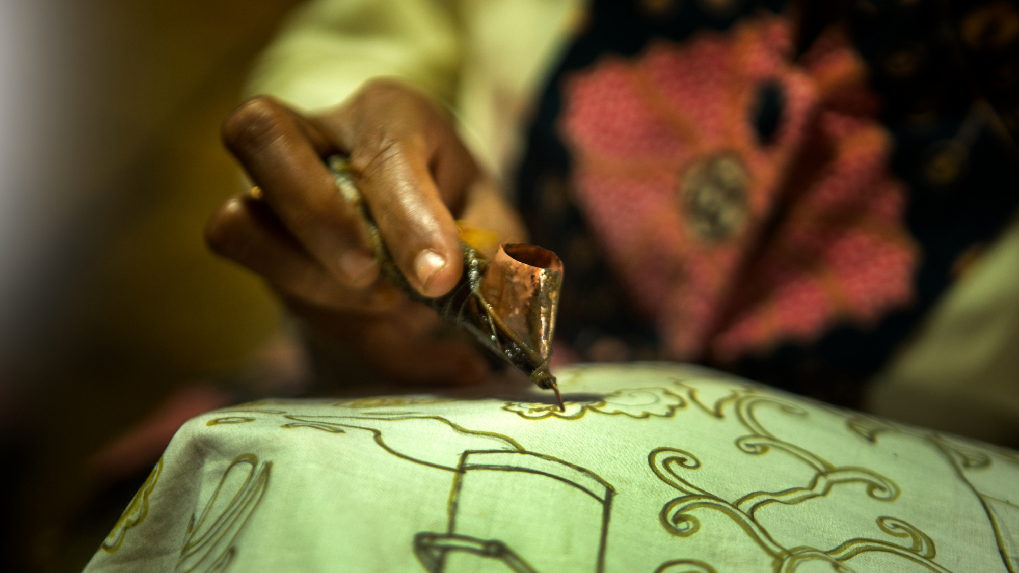 A person's hand holds a tool used to paint intricate designs on green fabric.